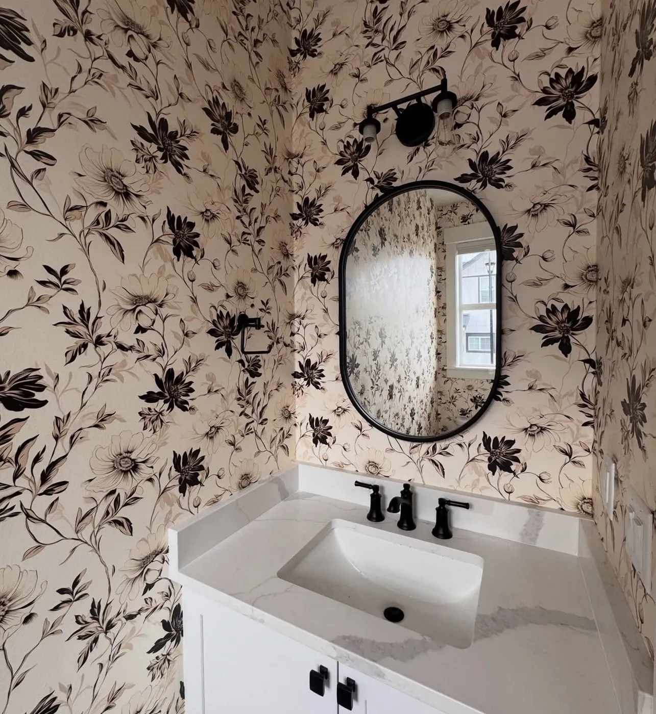 Powder room wrapped in black-and-cream floral wallpaper with an oval black mirror